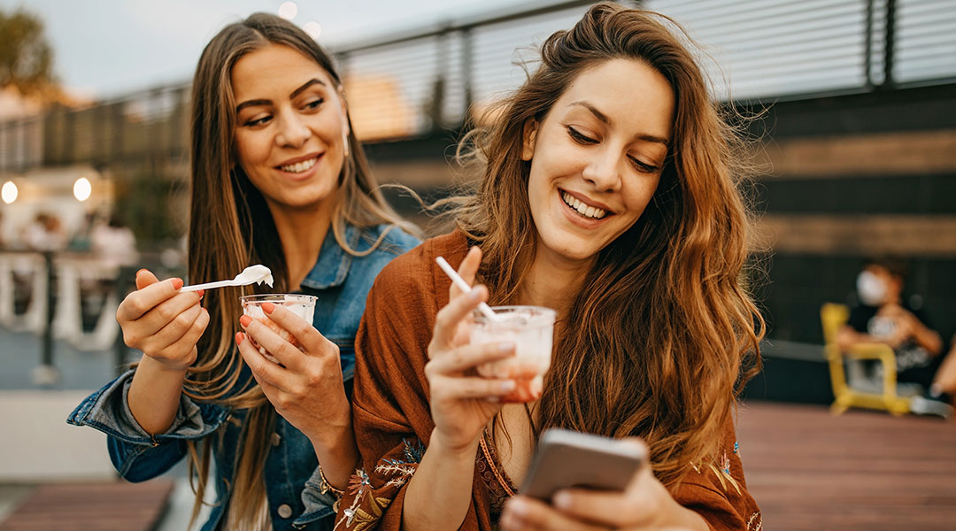 Two girls outdoors eat sundaes together. One is also looking down at her phone smiling.